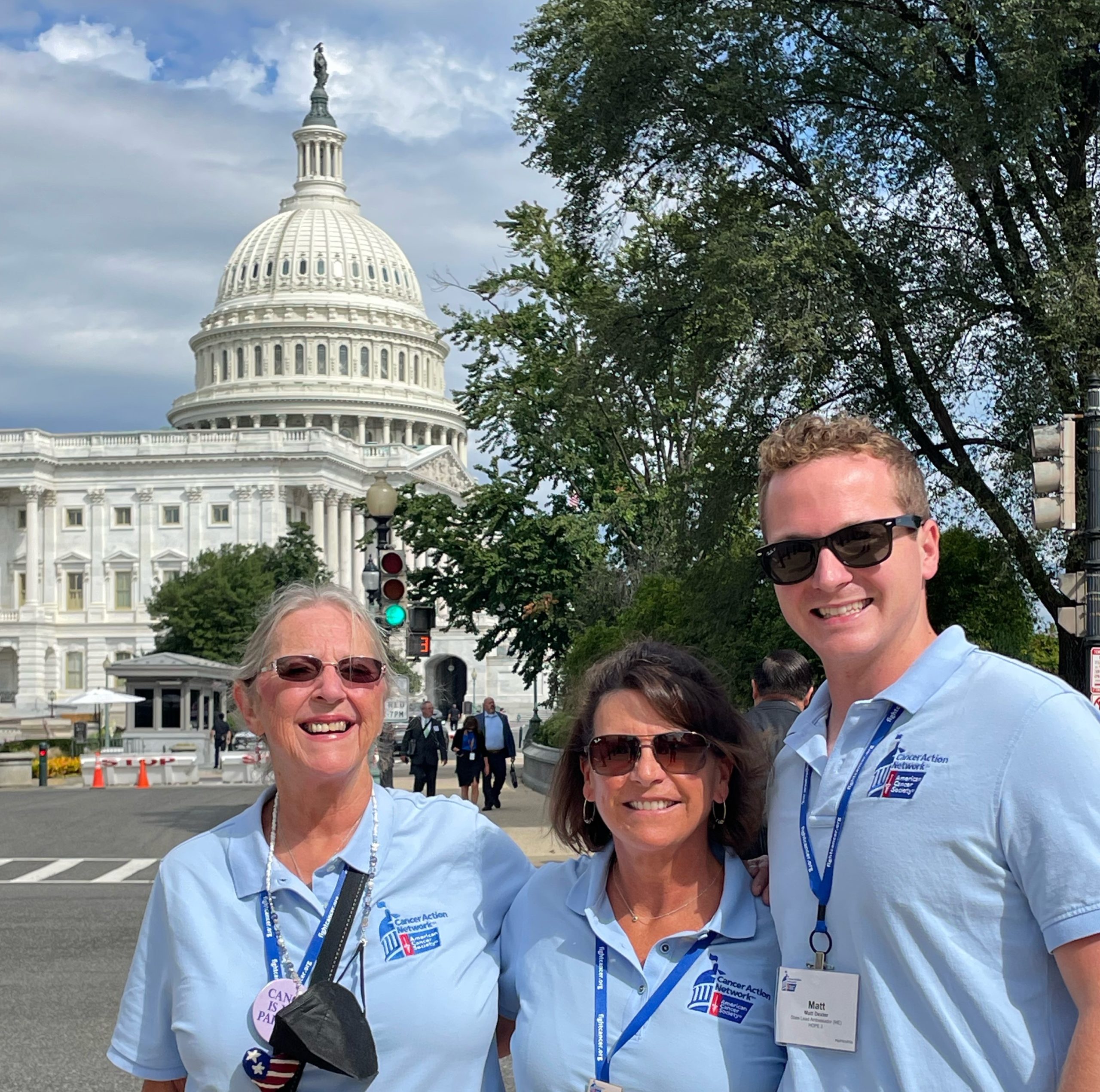 A group of three individuals, two women and a man, stand on a sidewalk with the U.S. Capitol building behind them.