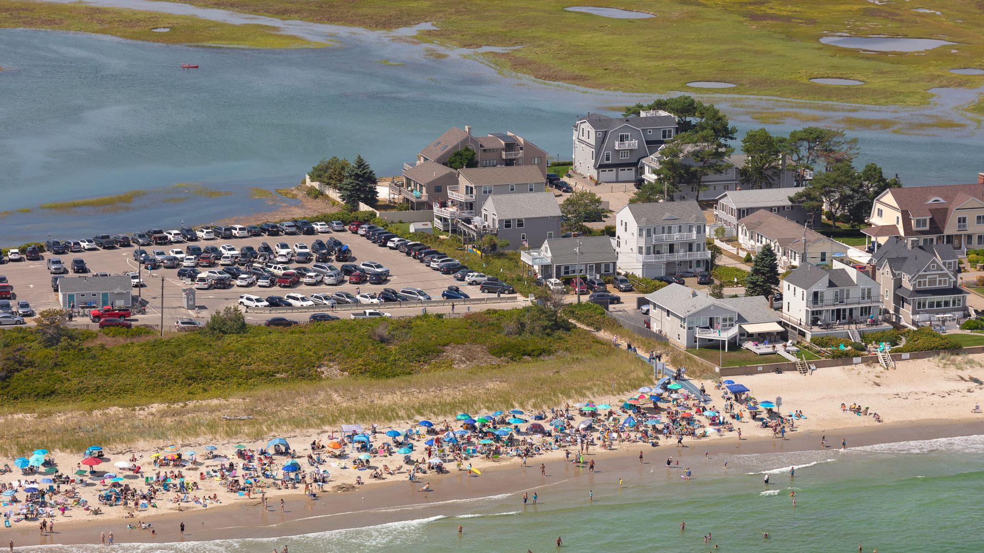 Aerial view of a beach in Wells filled with beachgoers. Behind the beach are rows of houses and a parking lot, all at risk of the surrounding water