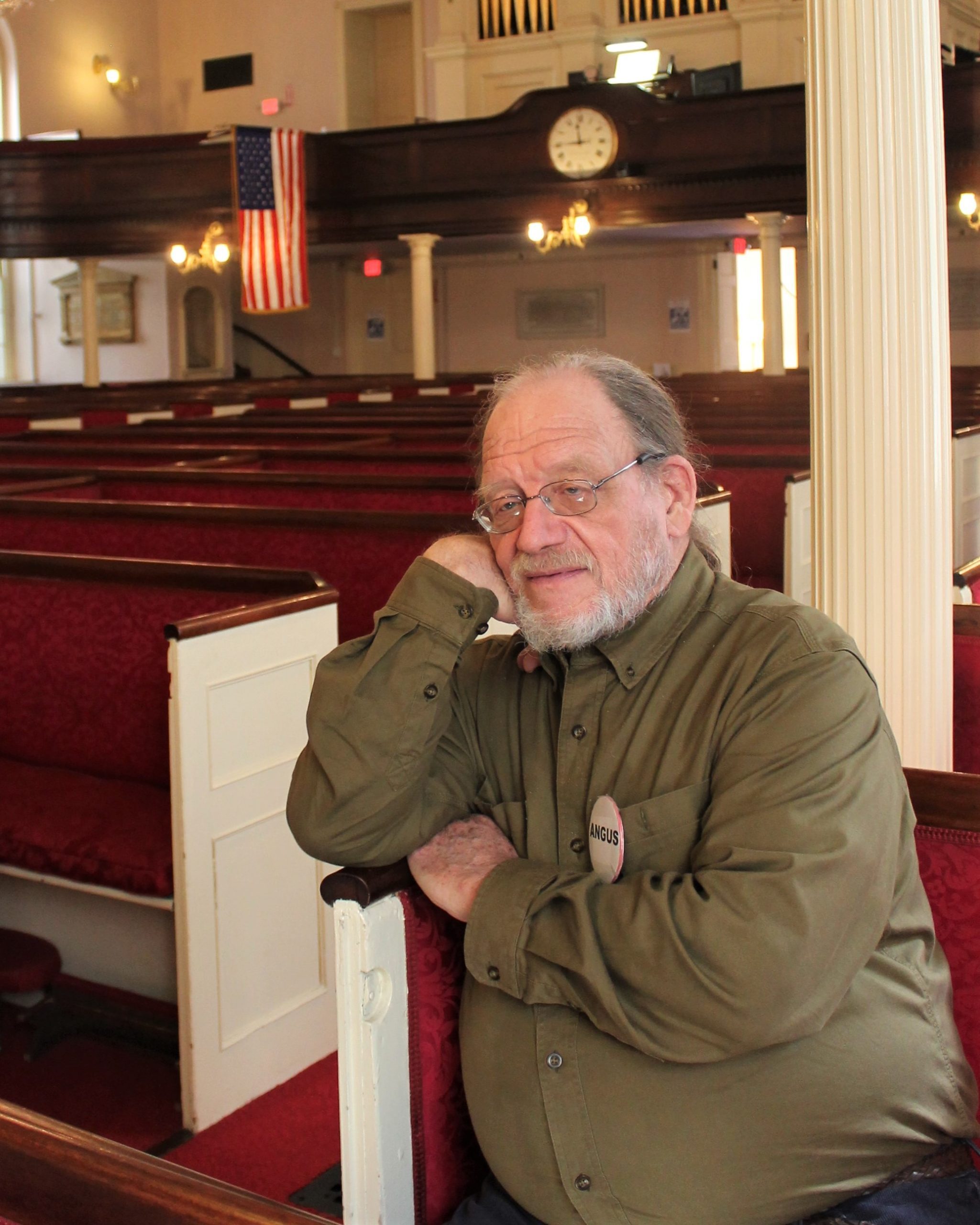 Angus Ferguson sits in one of many red church pews. 